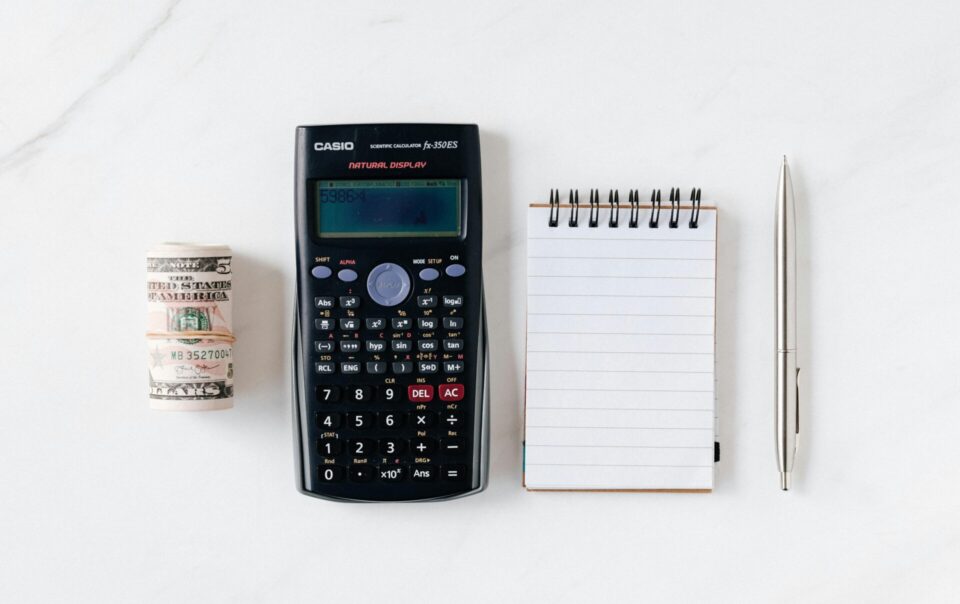 A roll of U.S. dollar bills, a Casio scientific calculator, a blank notepad, and a silver pen neatly arranged on a white marble surface as a basis for how to build a budget / simple budget.