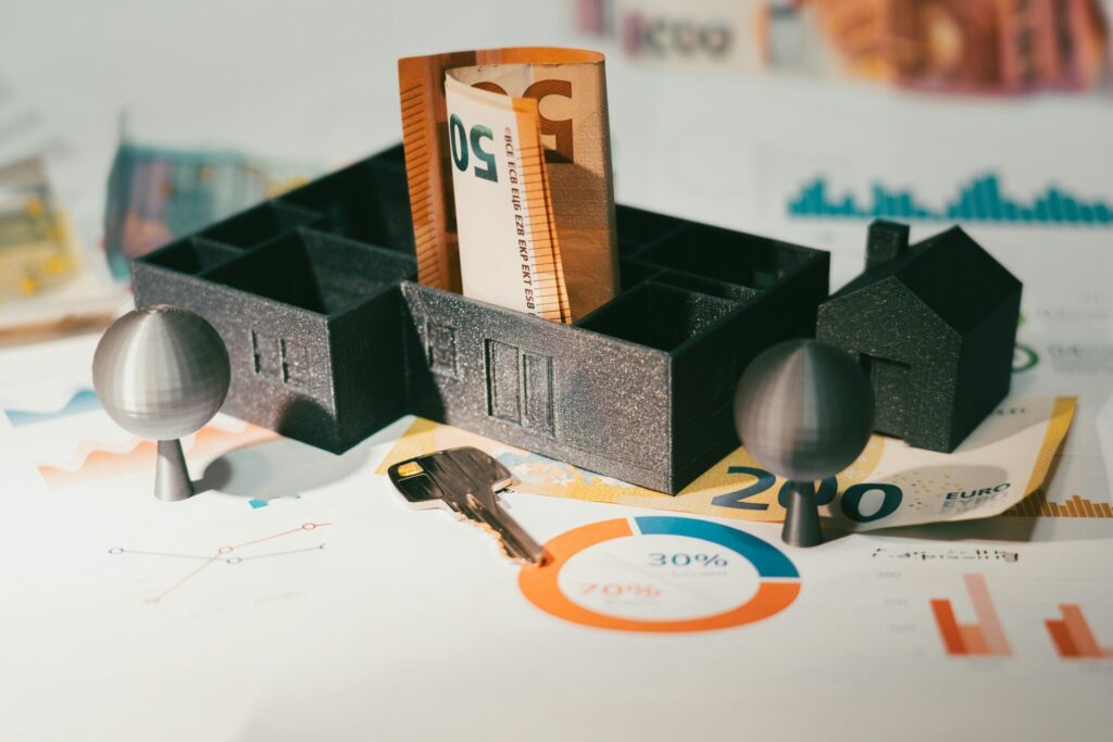 Model house with a rolled 50-euro bill inside, placed on documents with financial charts, a key, and euro banknotes- symbolizing real estate investment or mortgage planning.