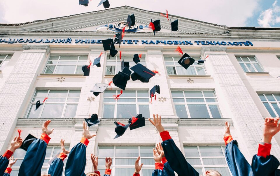 Graduates in blue gowns with red cuffs celebrate by tossing their black mortarboard caps into the air in front of a white university building with Cyrillic signage.
