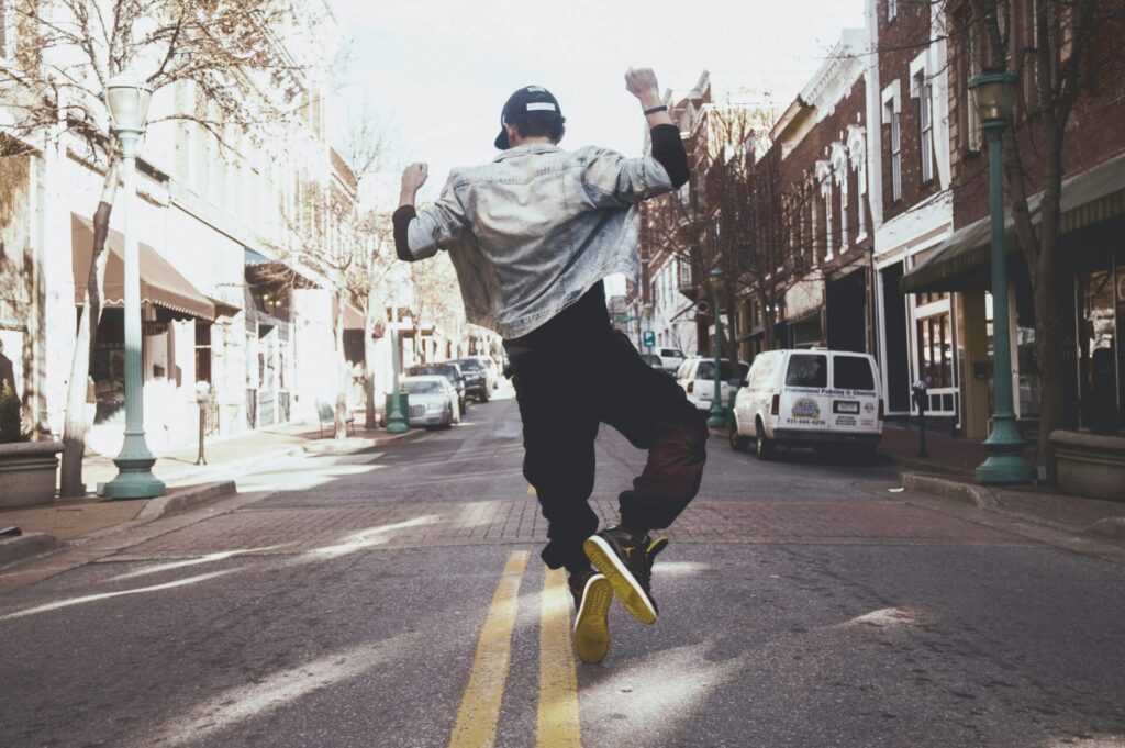 A young man wearing a denim jacket and a black cap jumps in the middle of a city street with his arms raised, surrounded by brick buildings, streetlights, and parked cars.