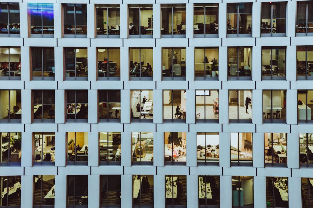 A modern office building with large windows, showing multiple floors with illuminated workspaces and people engaged in various activities inside.