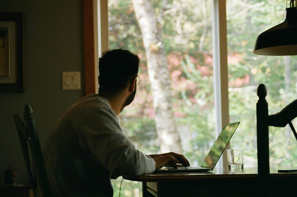 A man wearing glasses and a gray sweatshirt sits at a wooden desk with a laptop, gazing out of a large window at a green and slightly blurred outdoor landscape. The setting suggests a home office or a remote work environment with a calm and thoughtful atmosphere.