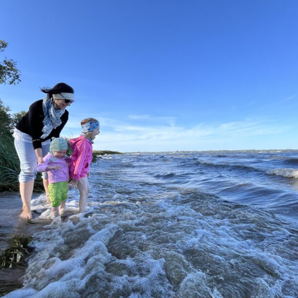 A woman and two young children play at the edge of a wavy seashore on a sunny day, standing barefoot in the water with grass and trees lining the beach in the background. Enabled by a precise line-by-line budget.