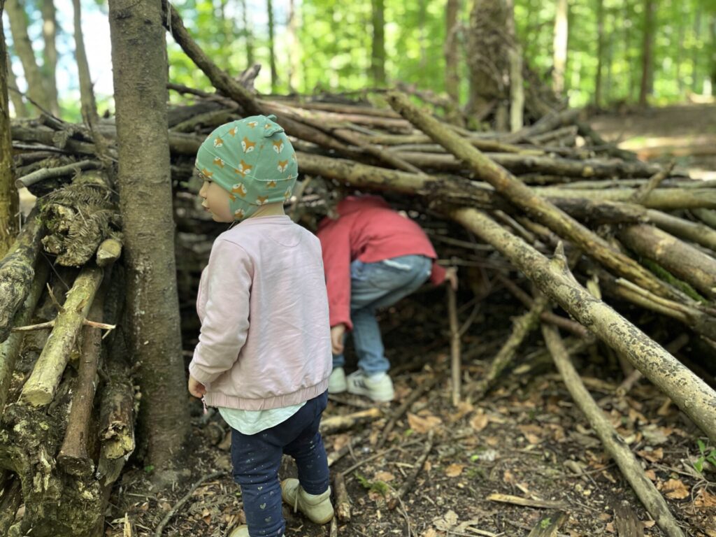 Two small children playing in a forest, exploring a handmade wooden hut constructed from large branches and logs. One child is standing outside in a green hat with foxes, while the other is crawling inside the structure.