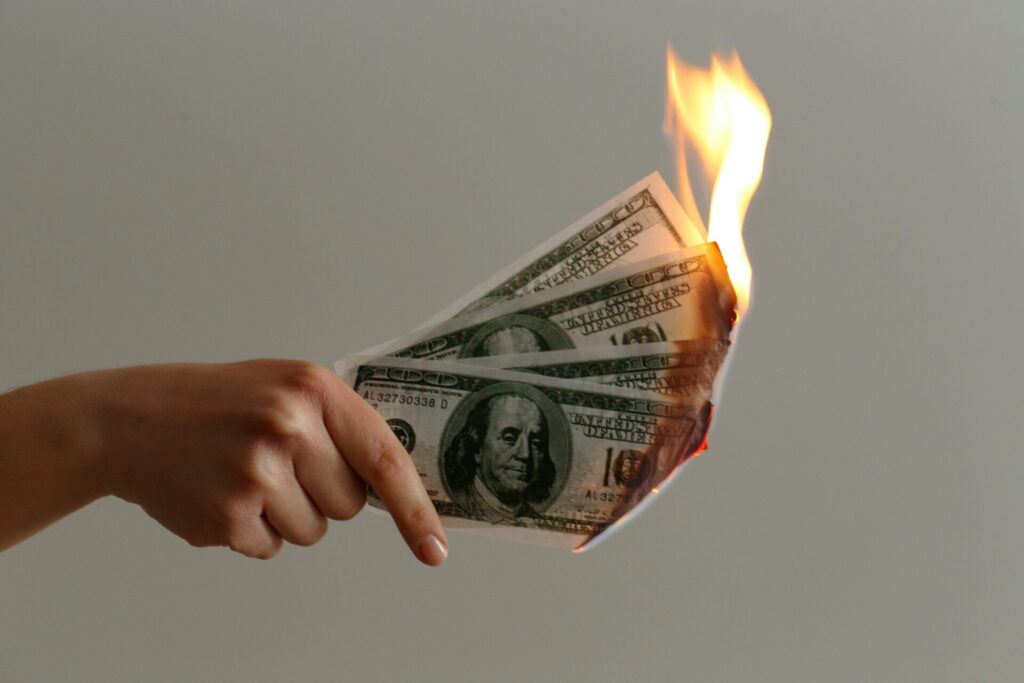 A hand holding burning U.S. hundred-dollar bills, symbolizing wasteful spending or financial loss.