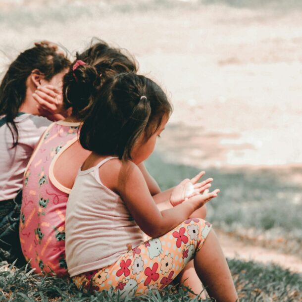 Three young girls sitting side by side on a grass lawn, viewed from behind — illustrating the joy and motivation behind pursuing financial independence with kids.